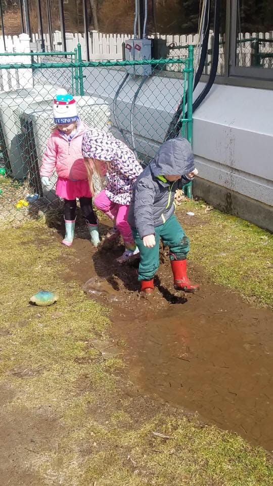 Three children playing in a muddy puddle outdoors