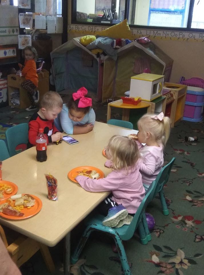 Children sitting at a table, some eating, some looking at a phone