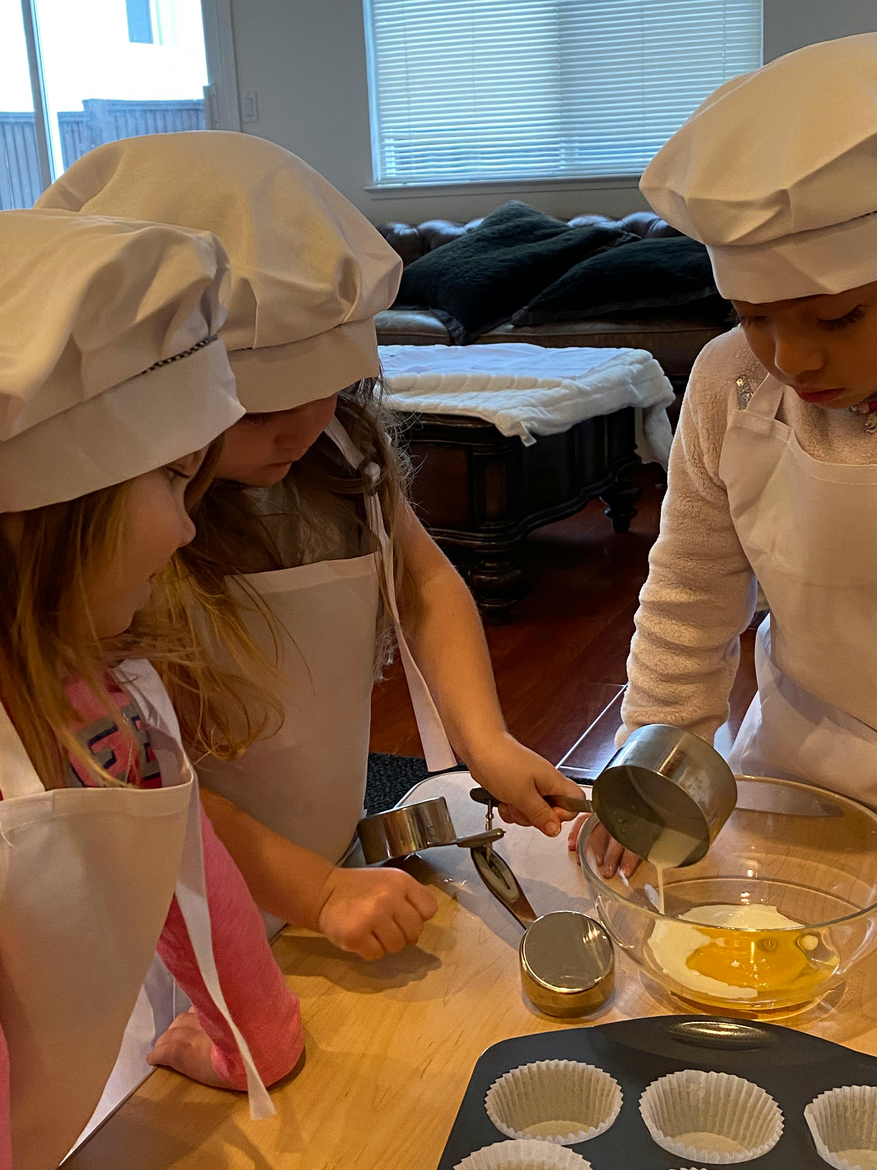 Three children in chef hats and aprons baking, pouring ingredients into a bowl
