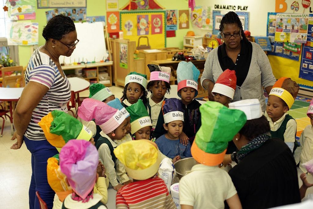 Two adults with a group of children wearing colorful chef hats in a classroom