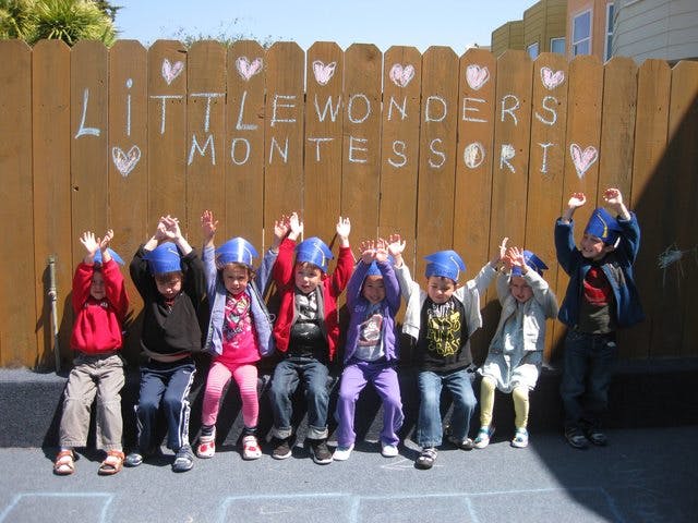 Children sitting in a row with hands raised in front of a wooden fence
