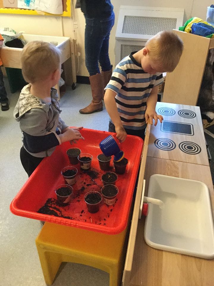 Two children playing with small pots of dirt in a red tray