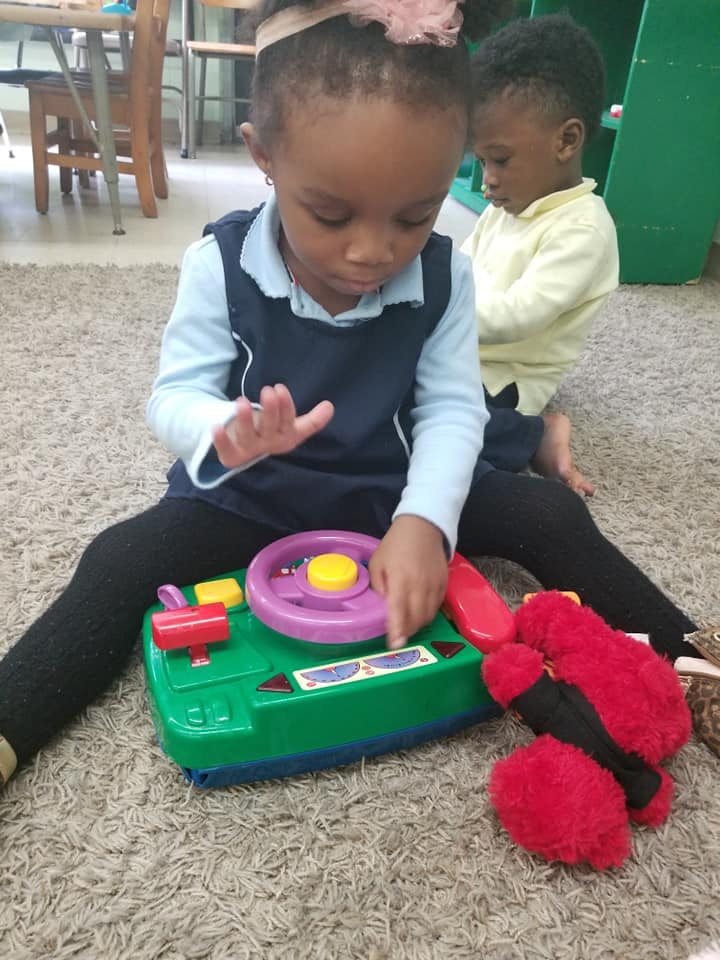 Child playing with a toy on the floor, another child in the background