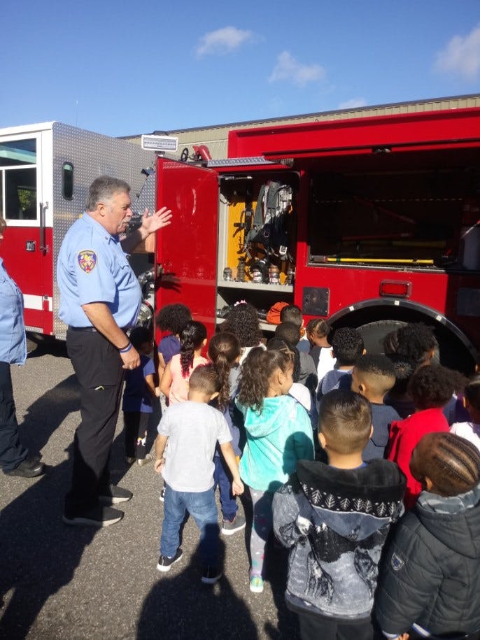 Person in uniform speaking to a group of children next to a fire truck