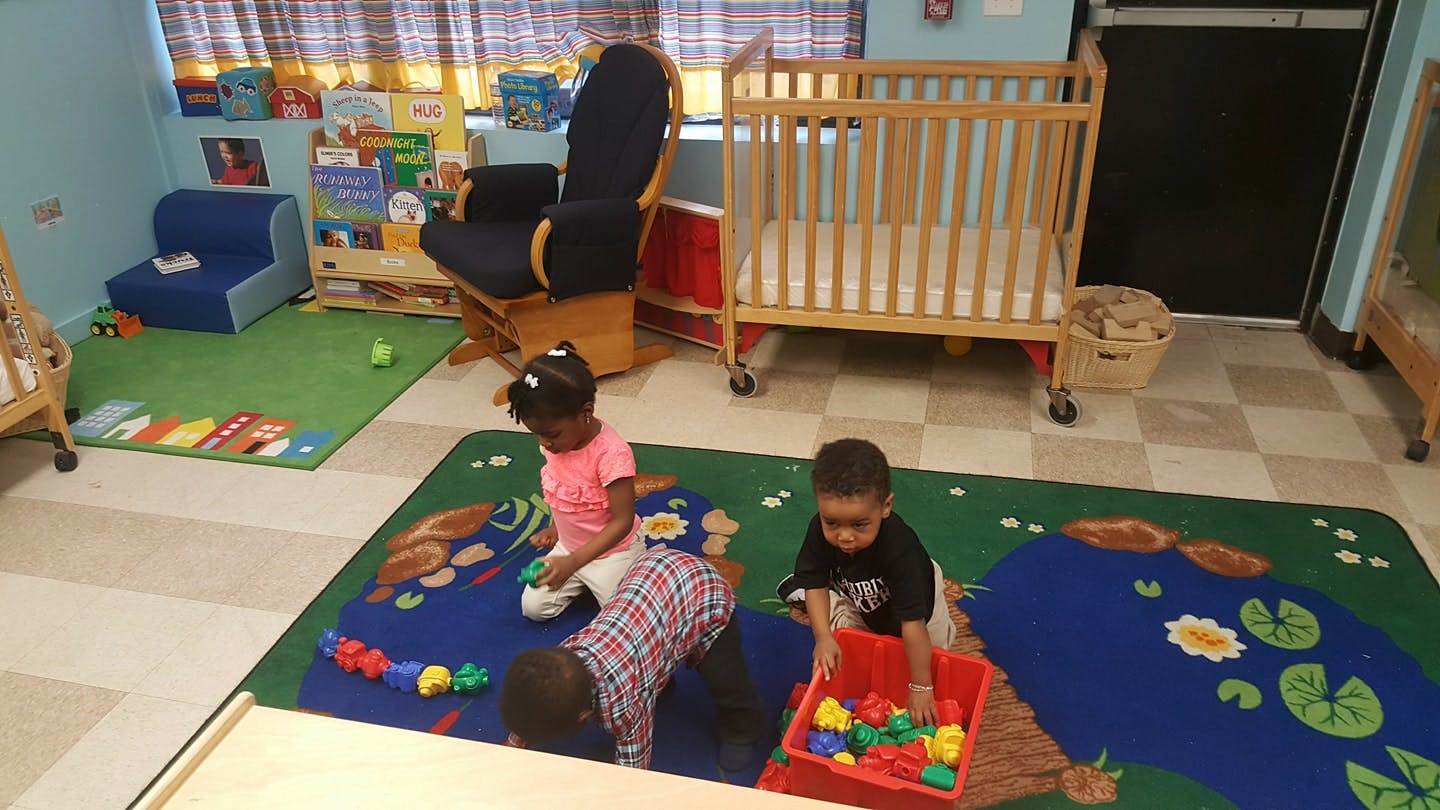 Three children playing with toys on a rug in a room with a crib in the background