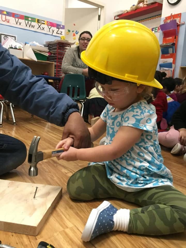 Child in a yellow hard hat holding a hammer with an adult