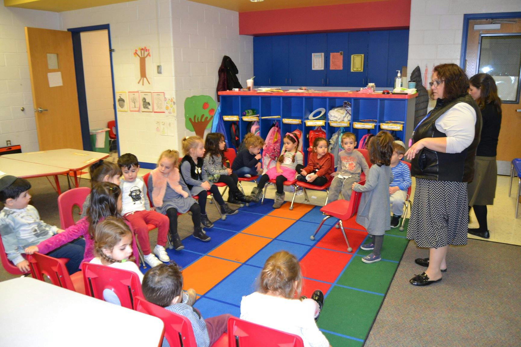 Adults and children in a classroom, some sitting on a colorful mat