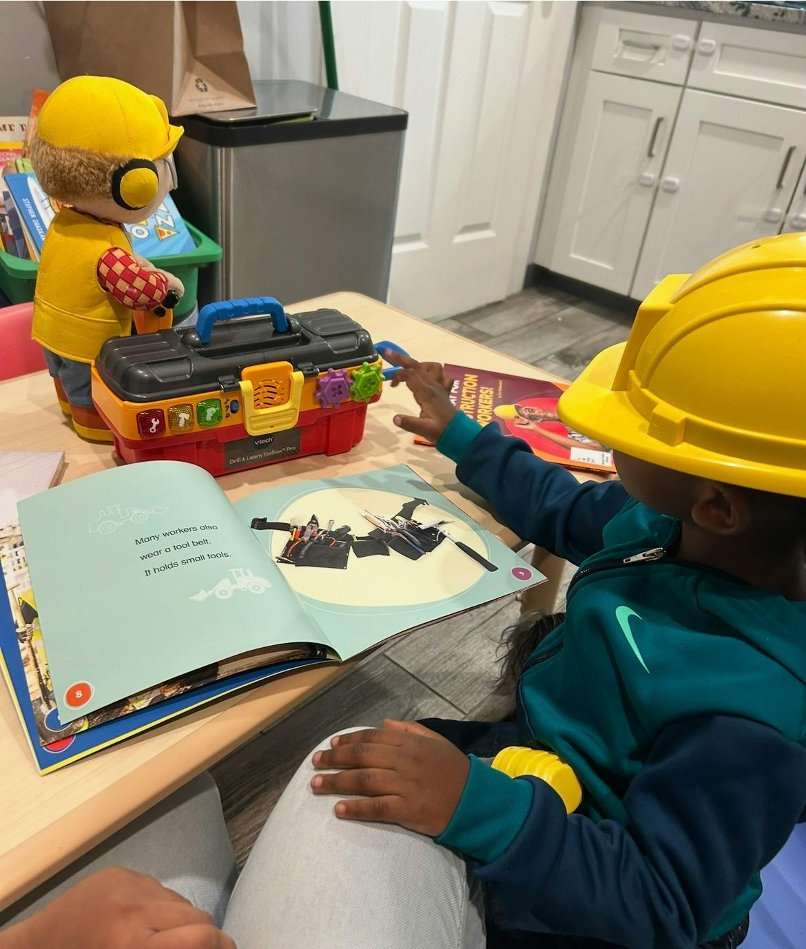 Child in a yellow hard hat pointing at an open book on a table