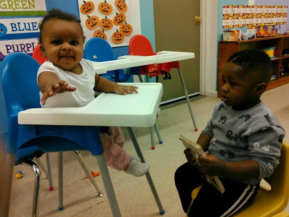 Child in high chair reaching out, another child sitting nearby