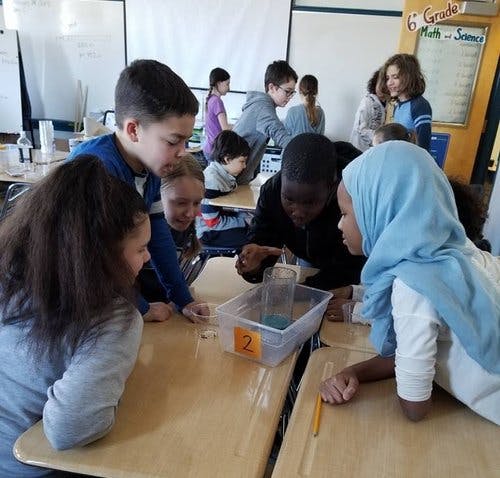 Group of children and teens gathered around a container on a desk in a classroom