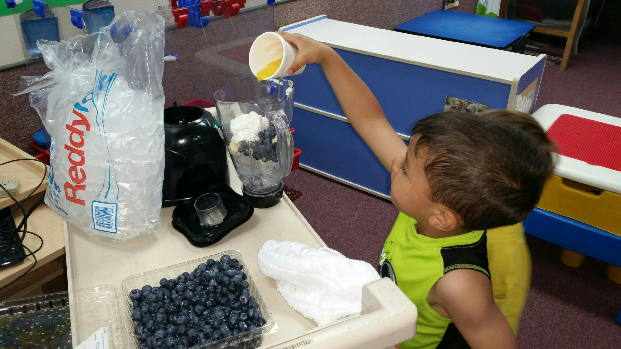 Child pouring liquid into a blender with blueberries and ice