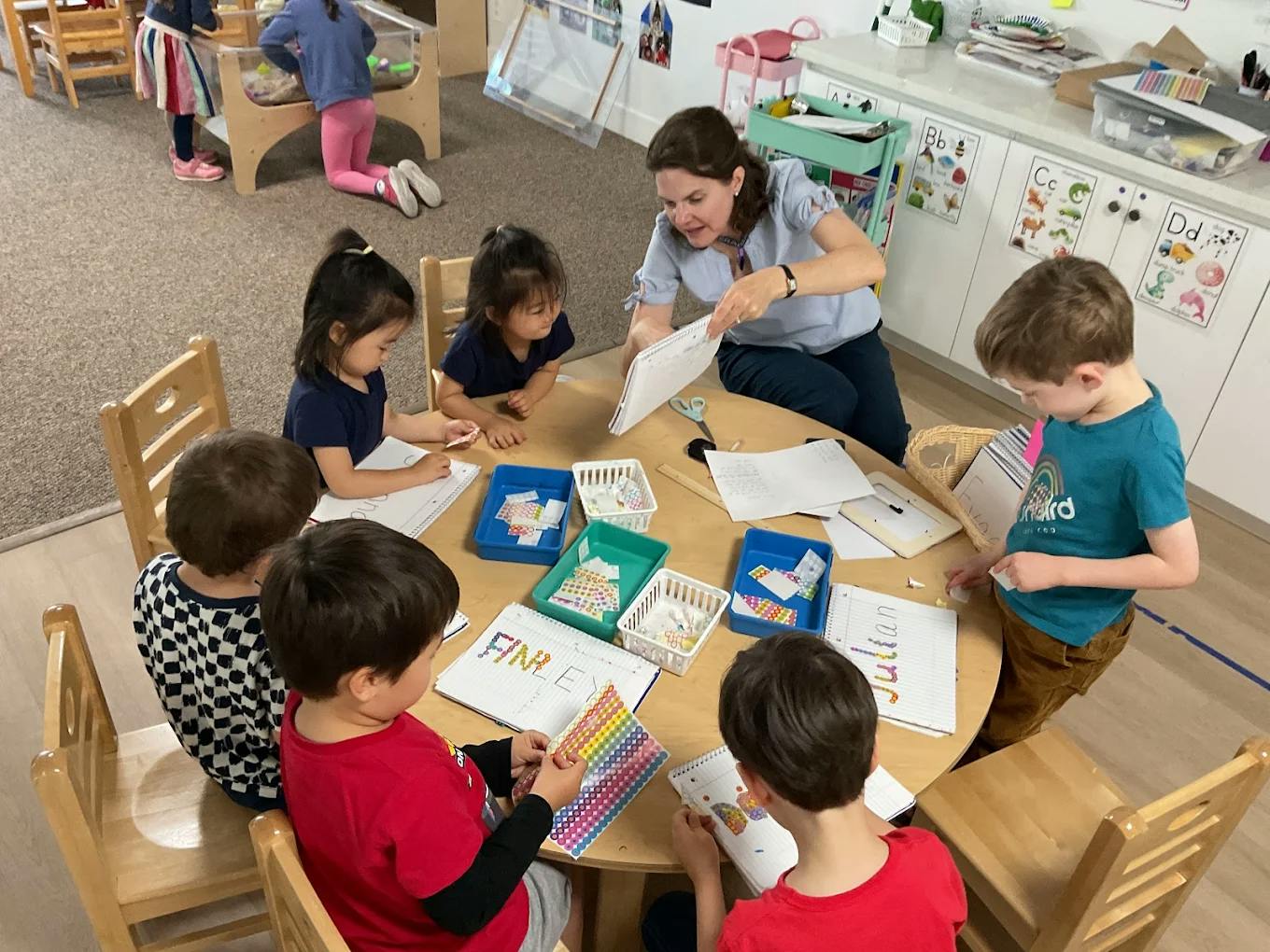 A person helping children with writing and stickers at a classroom table