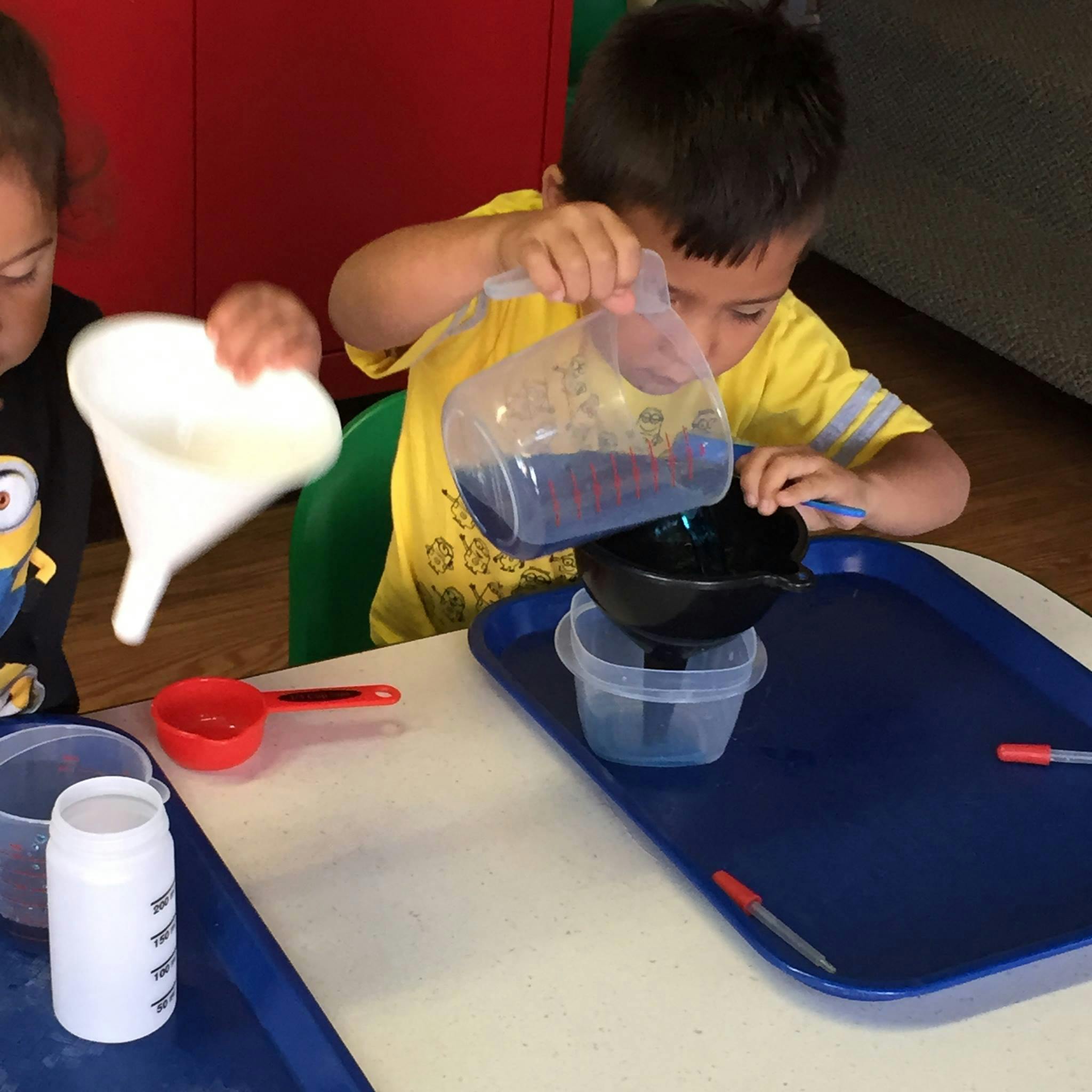 Two children pouring liquids into containers with funnels and containers