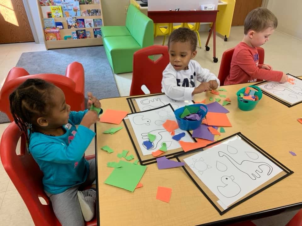 Three children sitting at a table doing crafts with paper cutouts