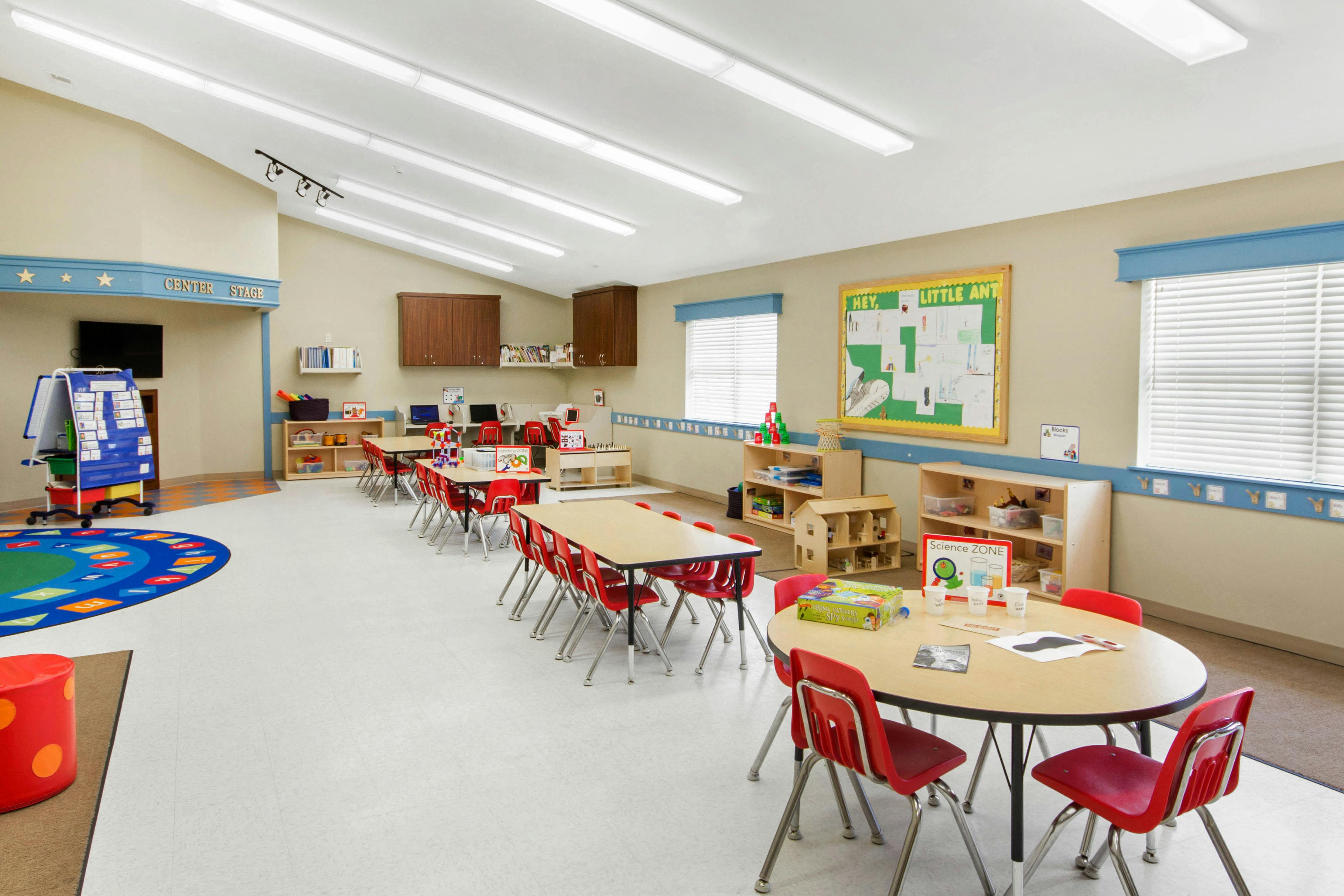 Classroom with multiple tables and red chairs, a rug with letters, and learning materials