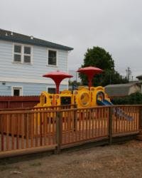 Playground with yellow train structure and red umbrellas behind a wooden fence