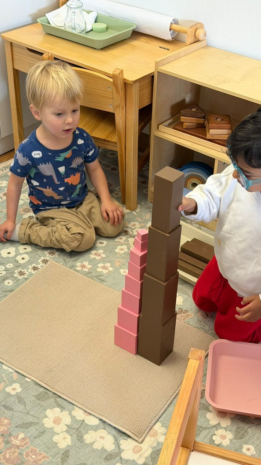 Two children on the floor, one building a tower of brown blocks, another watching