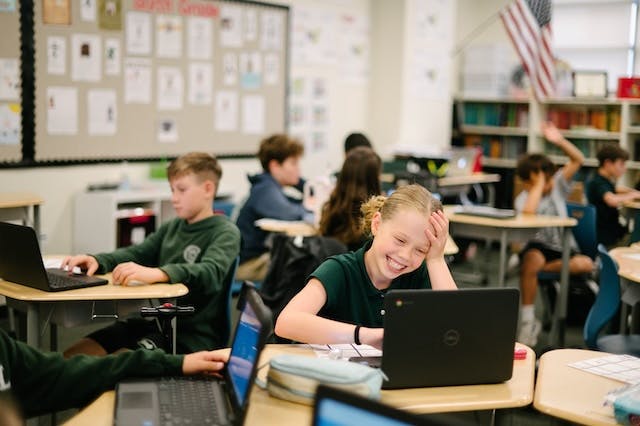 Child at a desk with a laptop, smiling with other children in a classroom