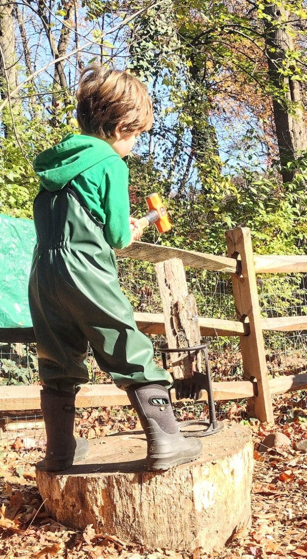Child in waders and boots standing on a tree stump holding a hammer