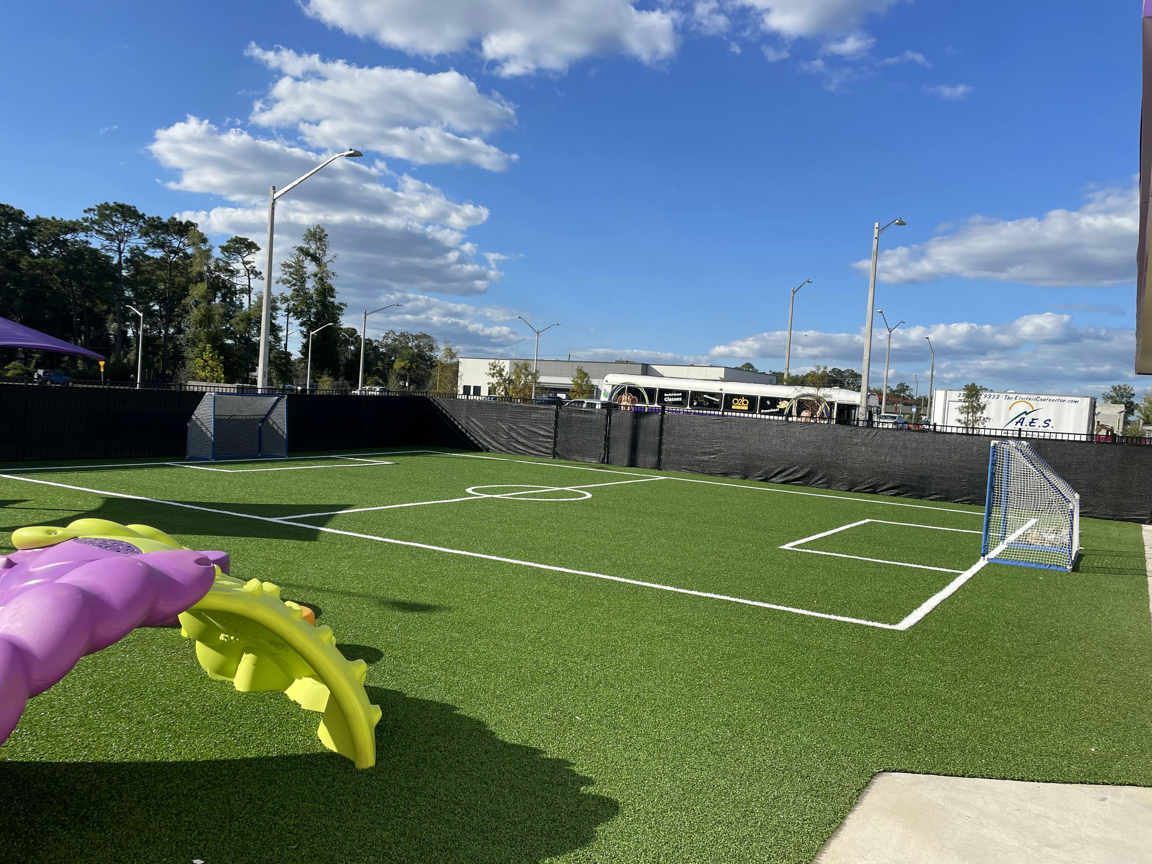 Fenced artificial turf play area with a small soccer goal and purple and yellow toy