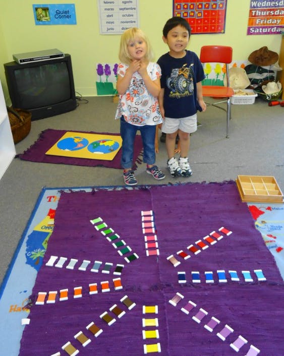 Two children standing in a classroom with colorful spools of thread on a rug
