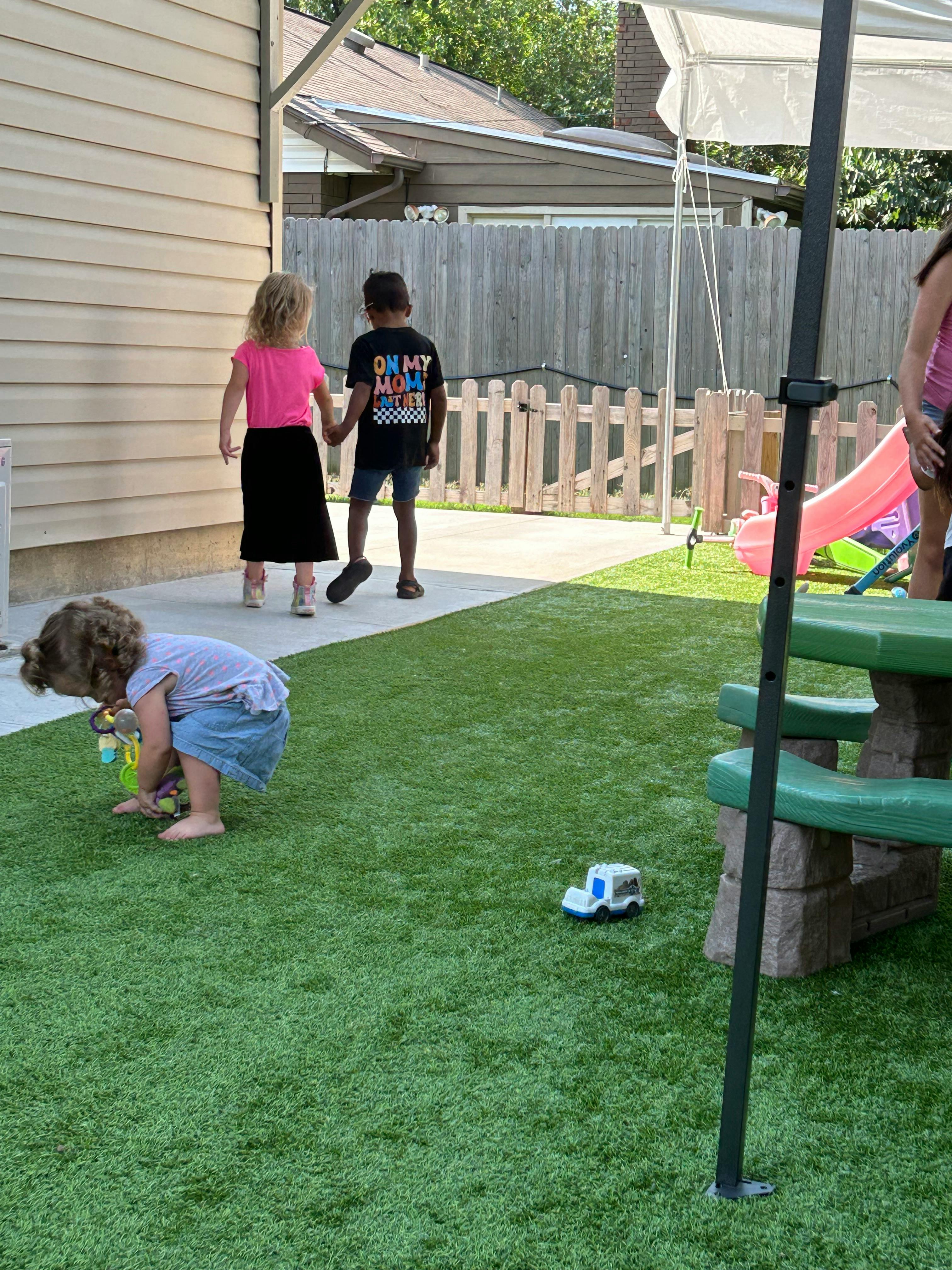 Two children holding hands walking away, another child playing on grass