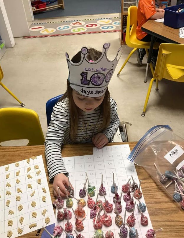 Child wearing a paper crown with "100 days smarter" written on it, counting items