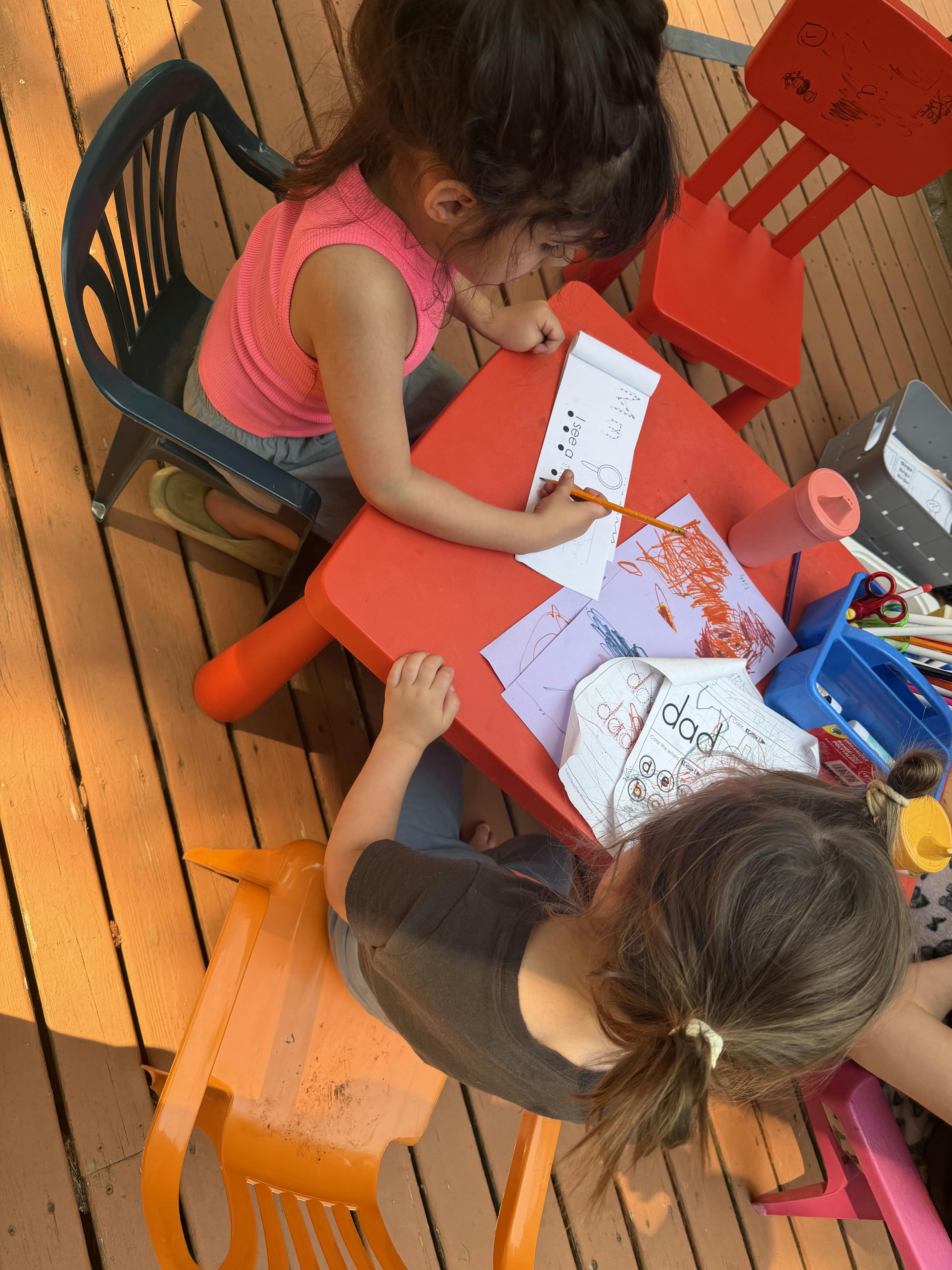 Two children sitting at a red table practicing handwriting and coloring