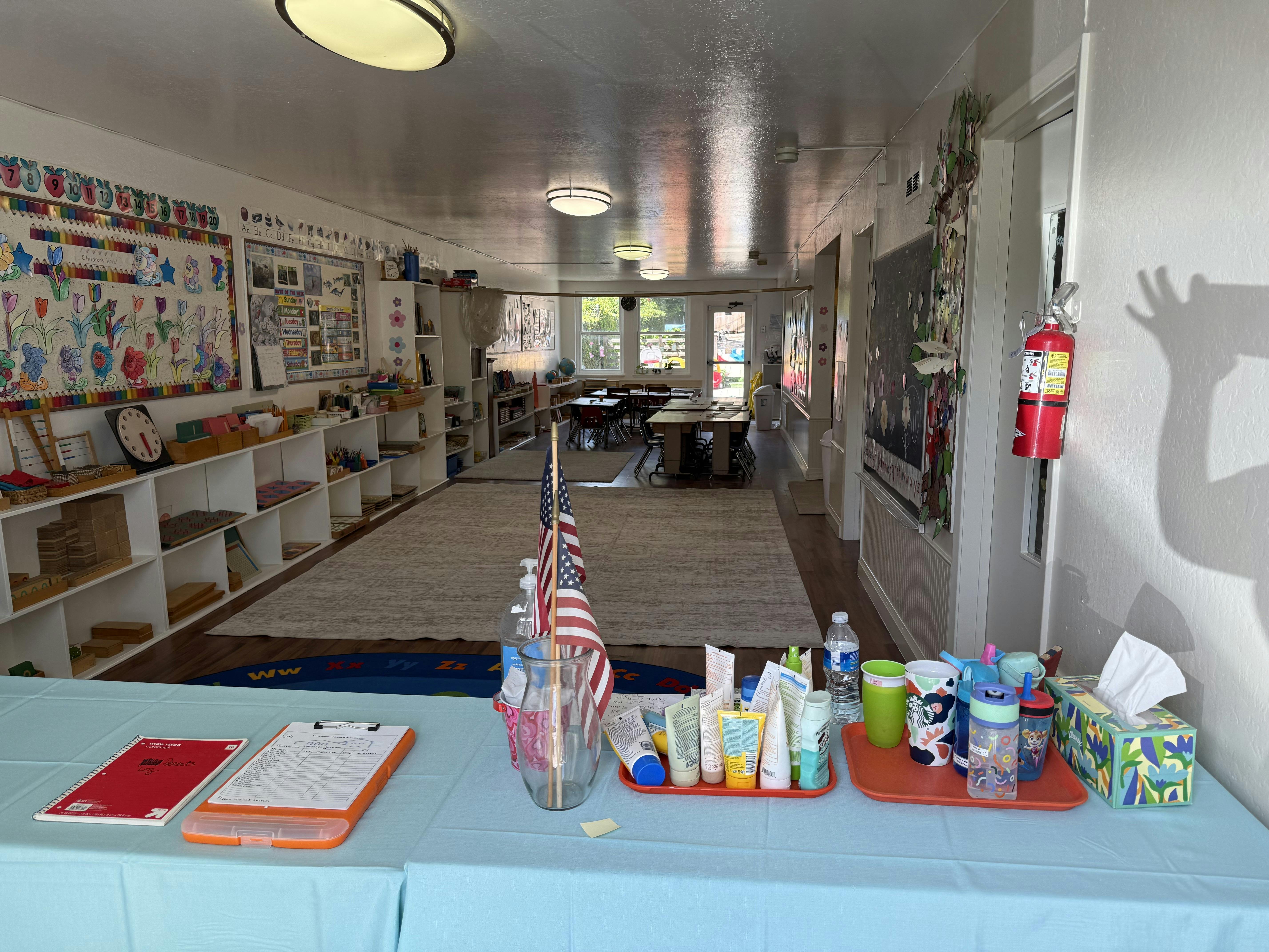 Classroom setup with shelves of educational materials, small tables and chairs, and a check-in table