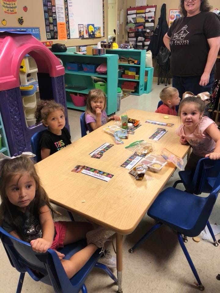 Children sitting at a table in a classroom with an adult standing nearby