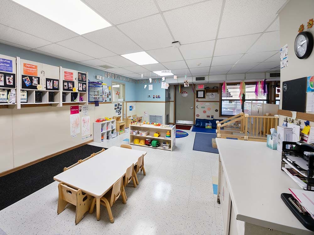 A brightly lit classroom with small tables and chairs, toys, and learning materials