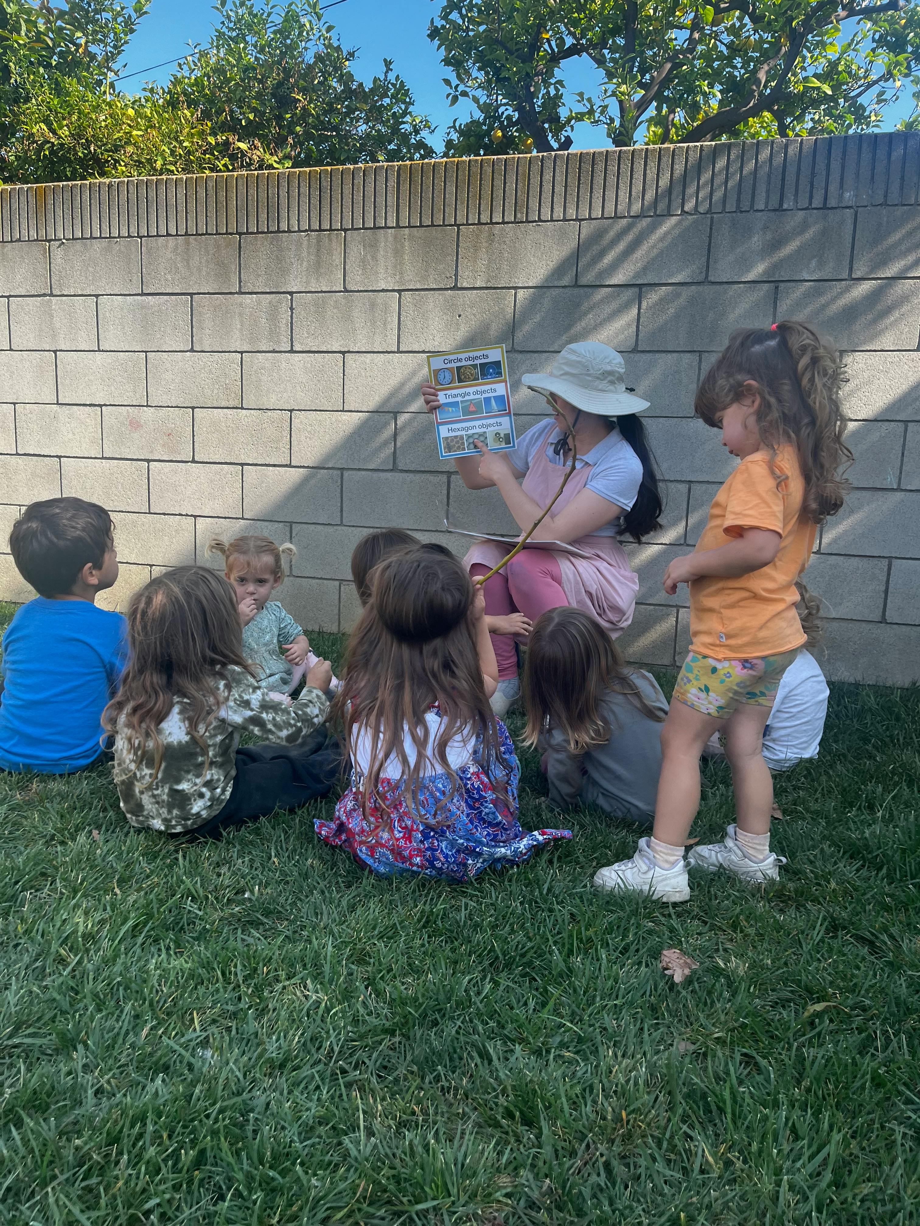 Person in hat reading a book to a group of children sitting on grass