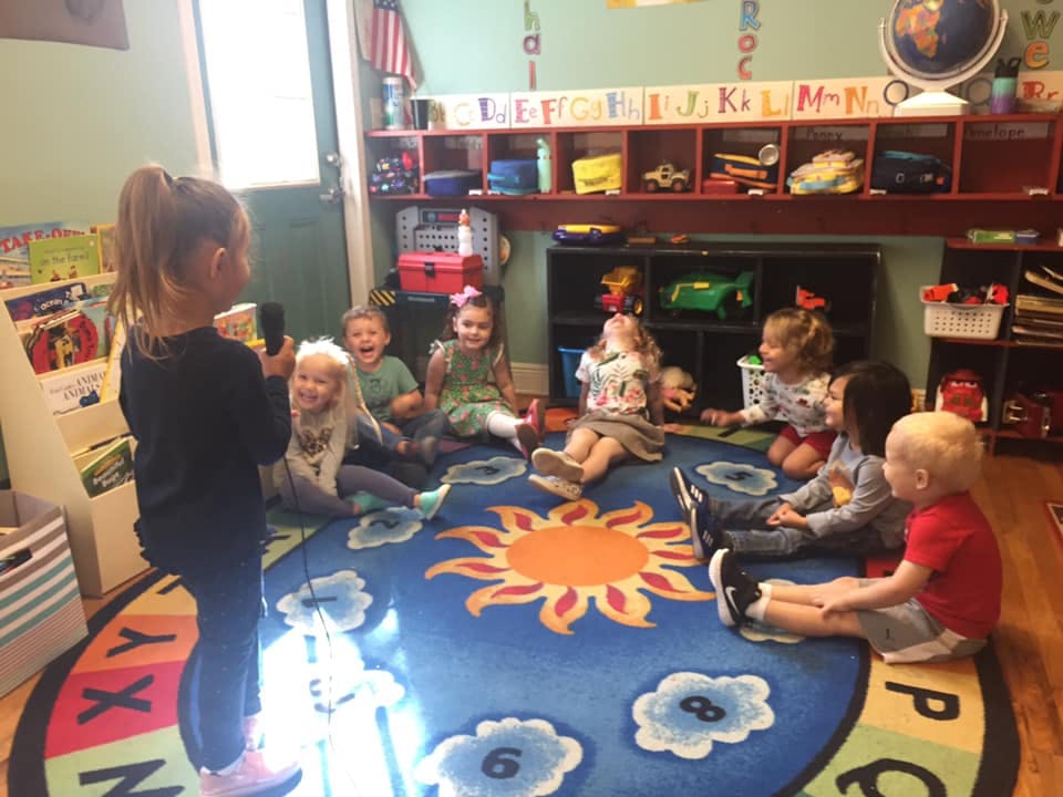 Child standing and speaking to a group of children sitting on a rug in a classroom