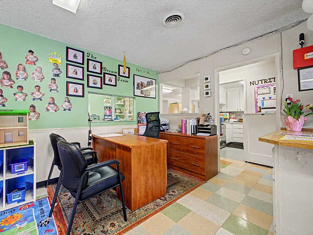 an office with a desk and chairs, a wall with children's photos, and a kitchen view