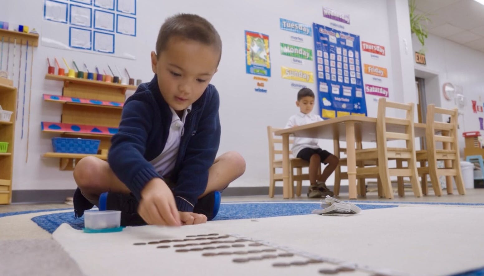 Child on floor arranging items, another child at a table in a classroom