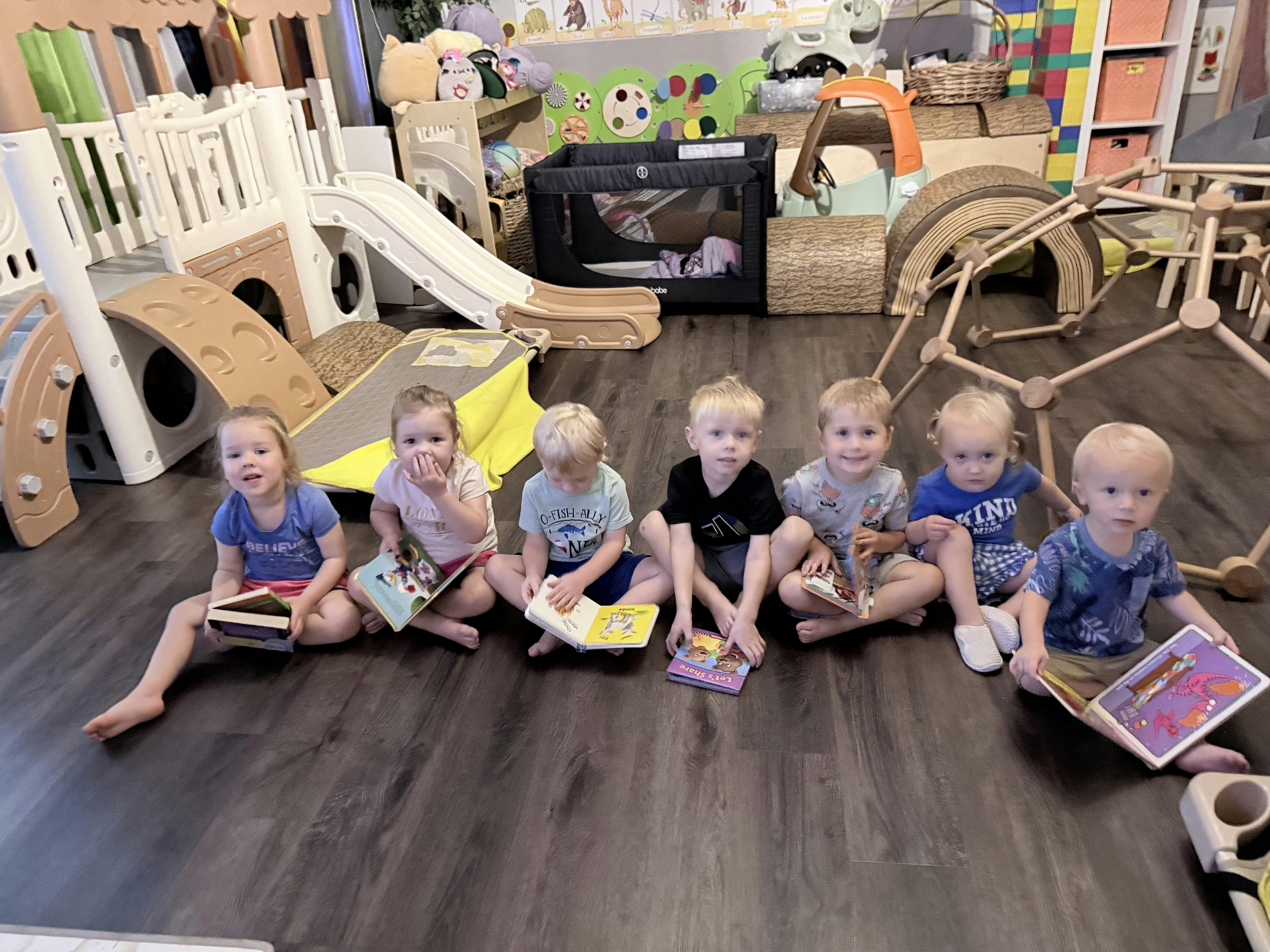 Seven children sitting in a row on the floor reading books in a playroom