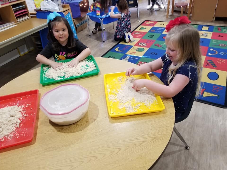 Two children playing with sand on trays at a table in a classroom
