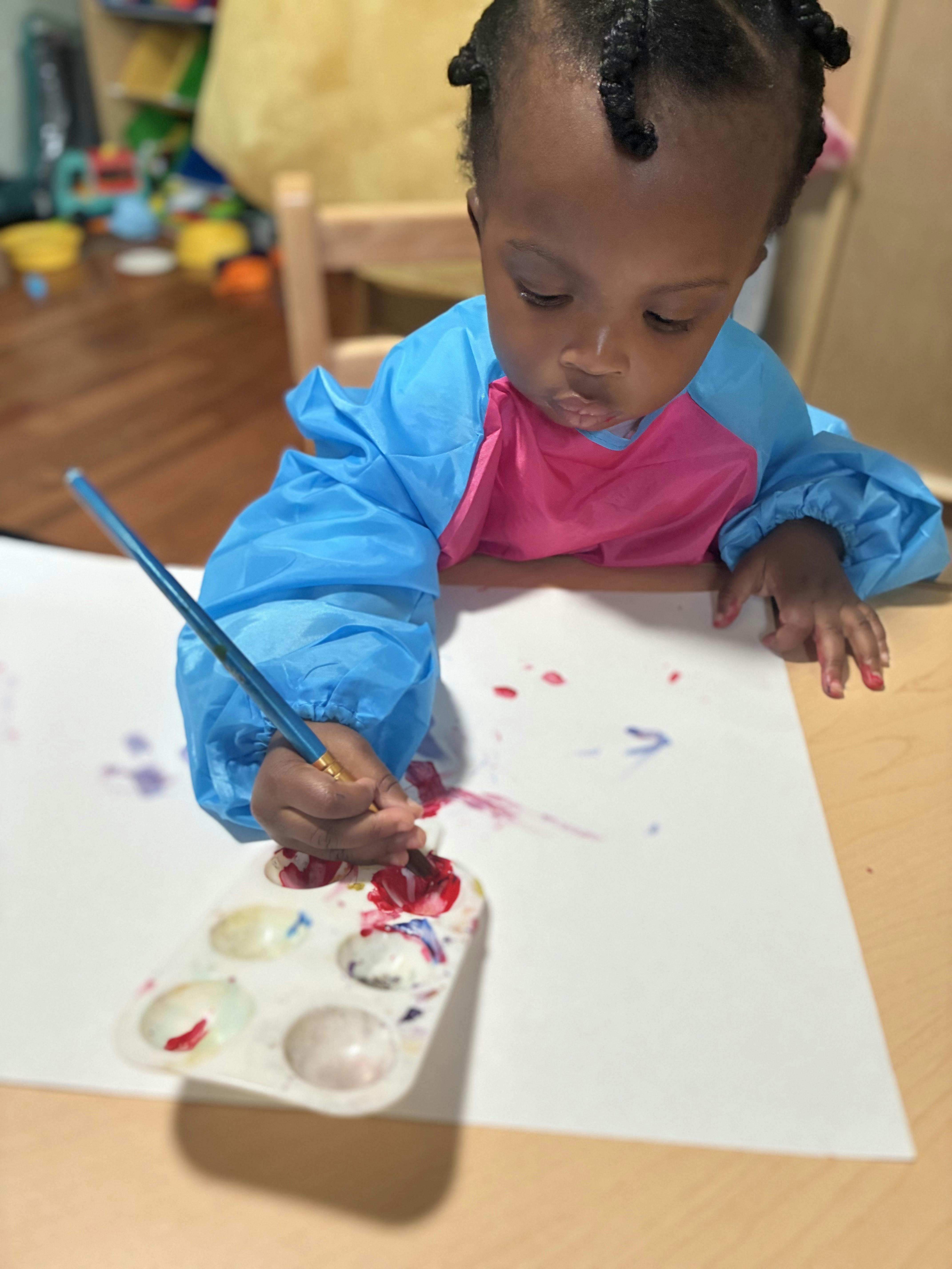Child in a smock painting at a table with a palette and paintbrush
