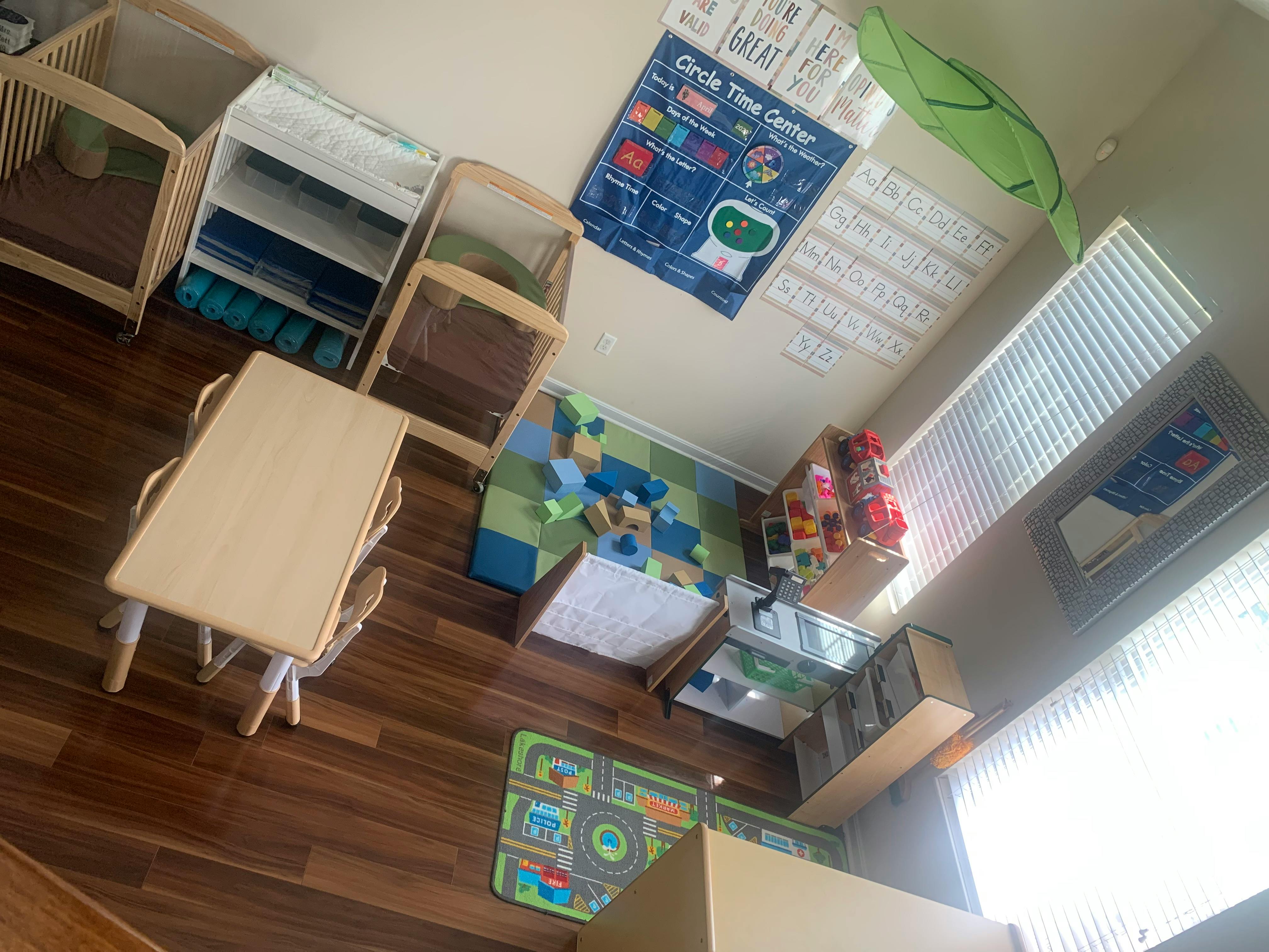 A classroom with small tables, chairs, cribs, and play mats