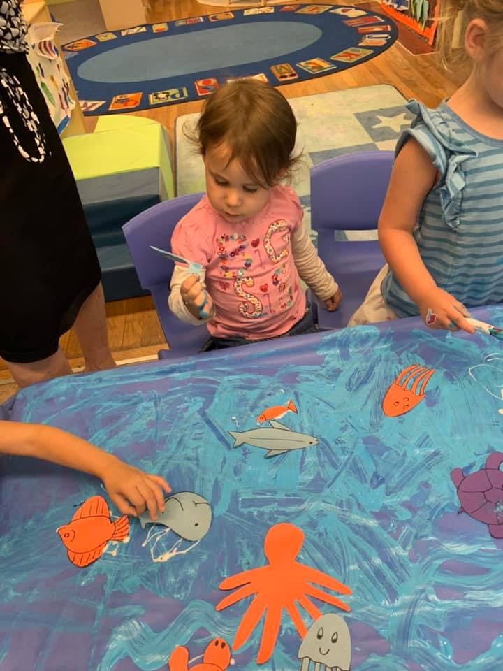 Child holding a paintbrush over a table with paper cutouts of sea creatures