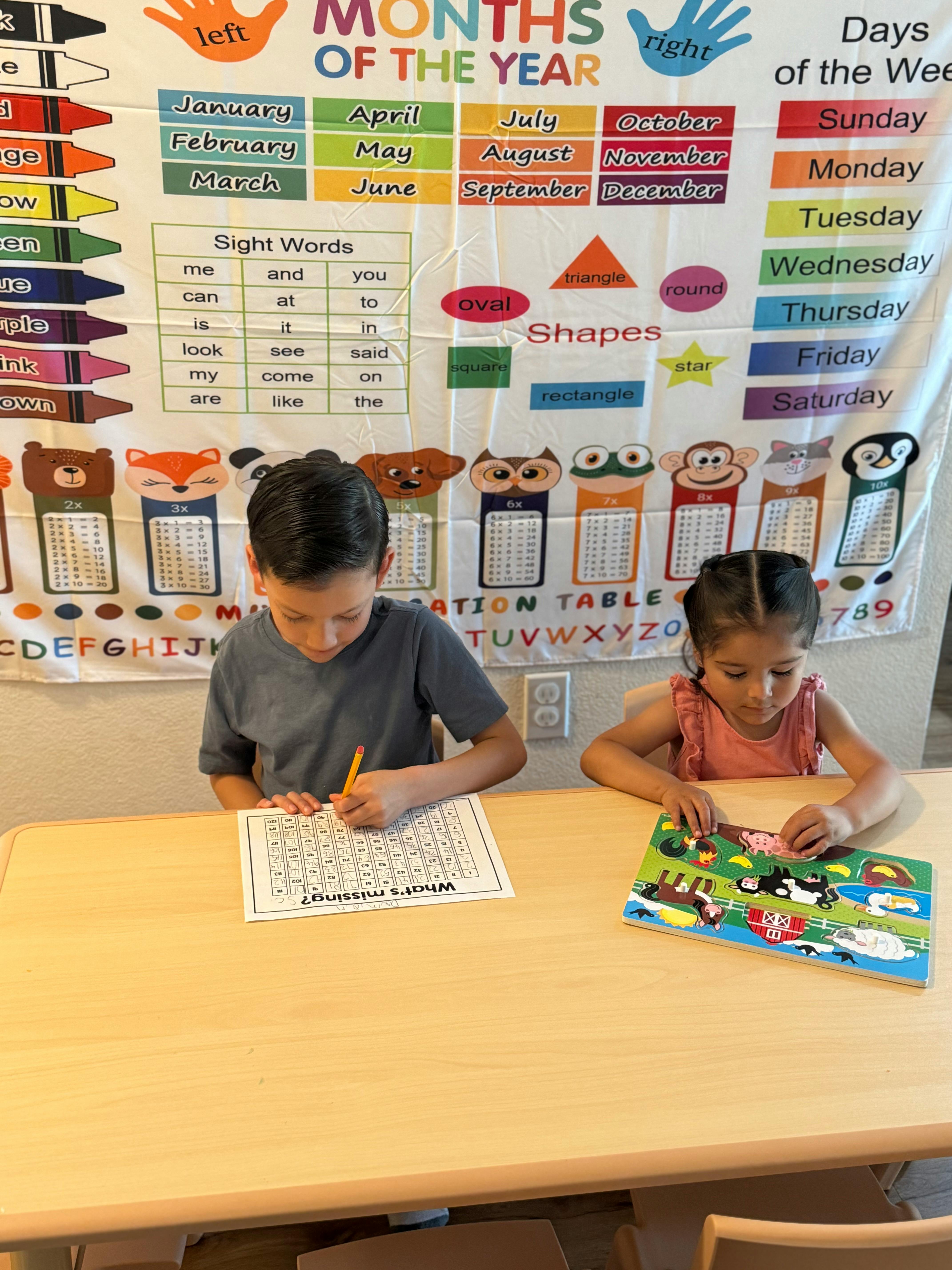 Two children at a table, one writing and one playing with a puzzle