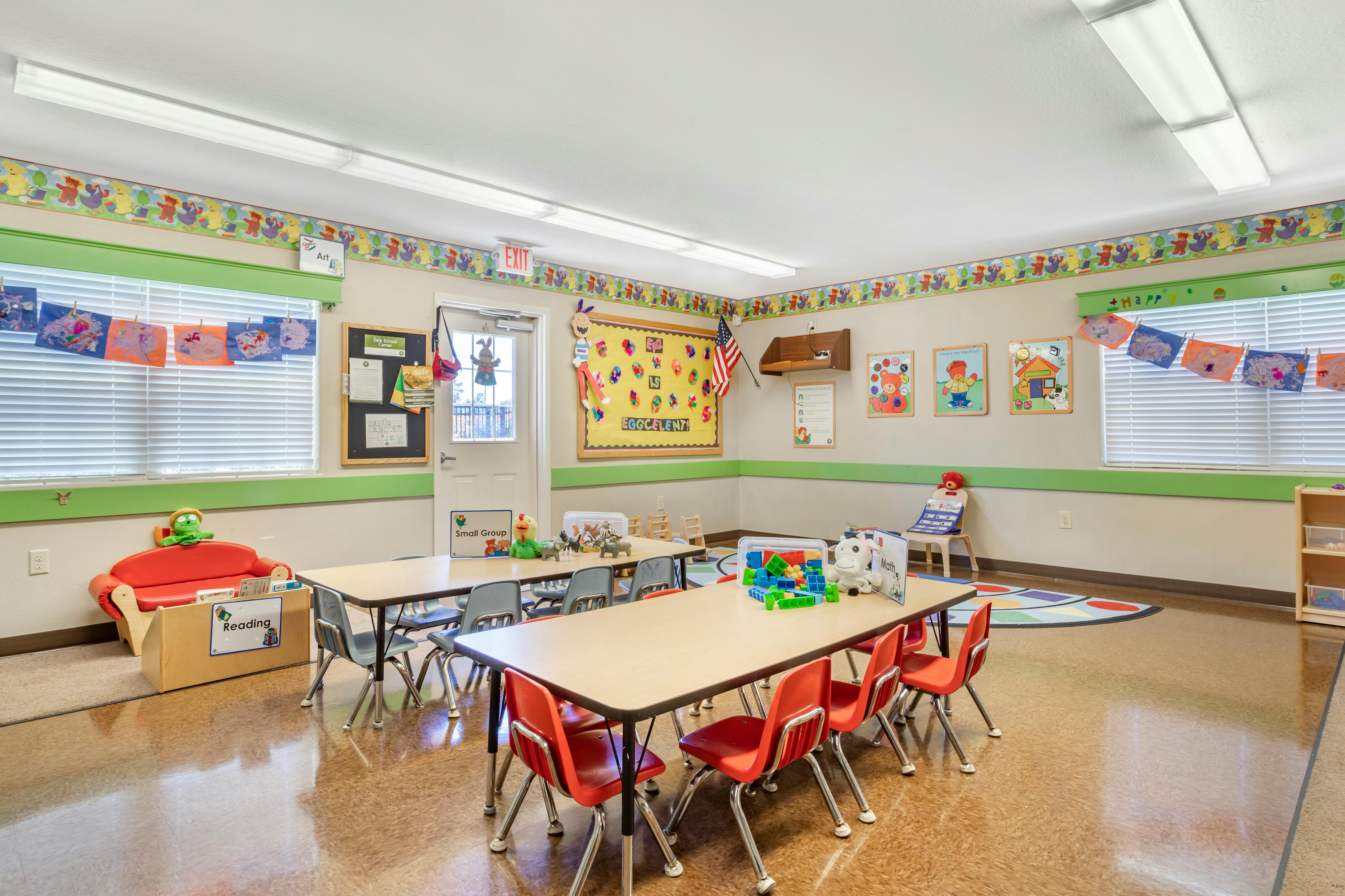 Empty classroom with small tables and chairs, children's artwork on walls and window