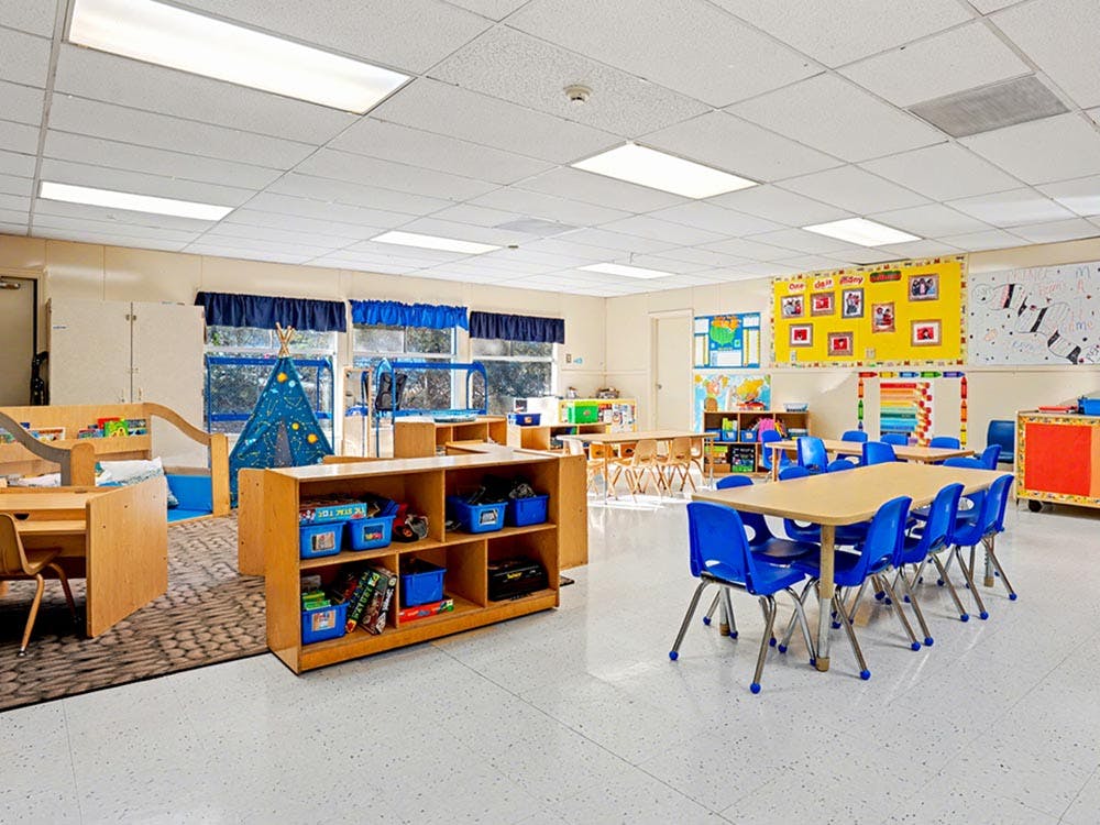 Classroom with small tables and chairs, a blue play tent, and shelving