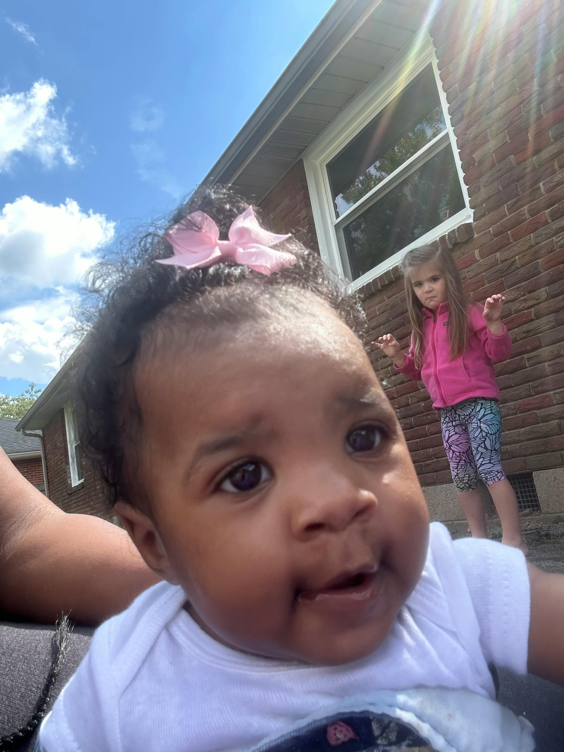 Close-up of a baby with a pink bow, another child in the background
