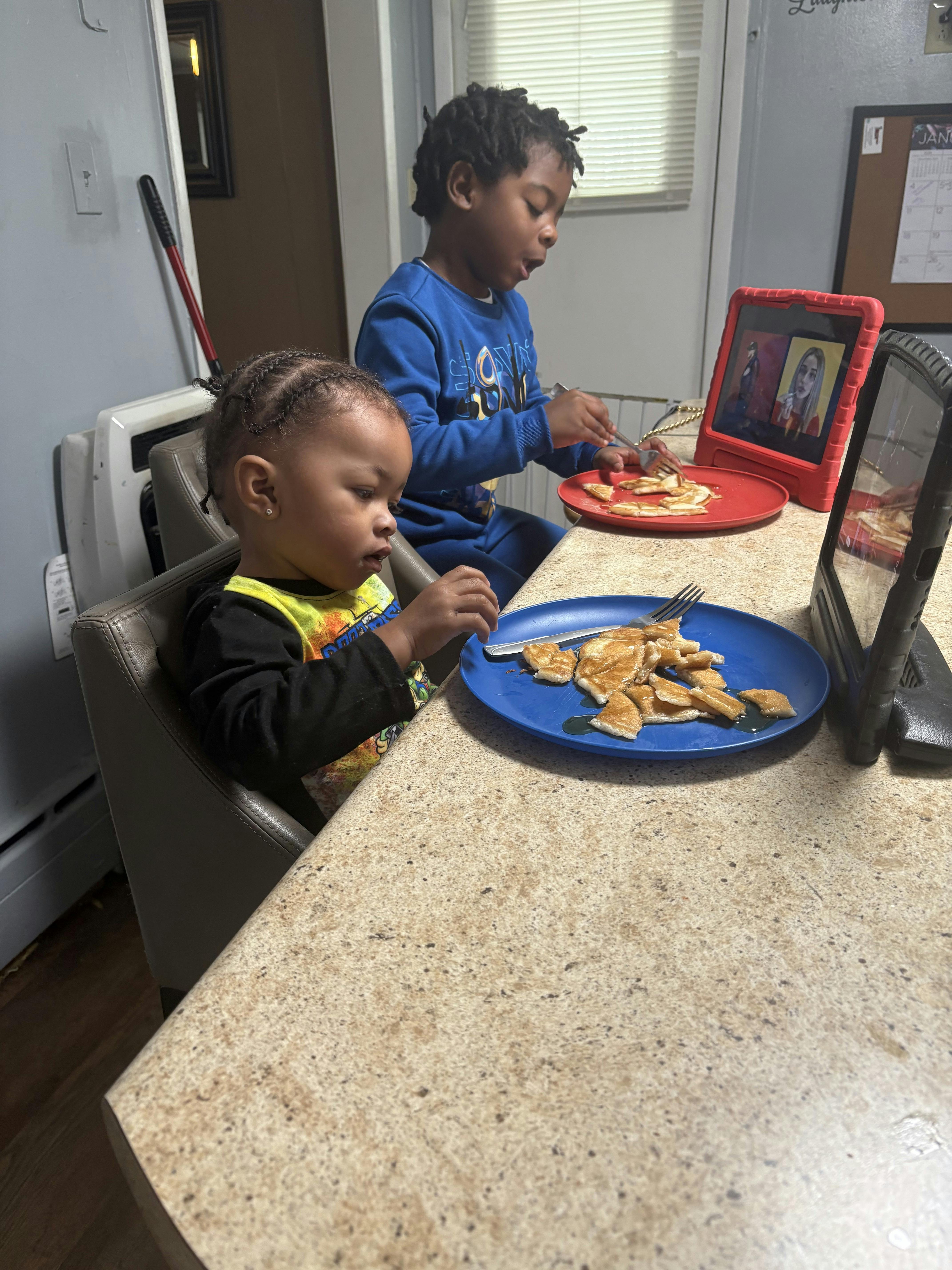 Two children at a counter eating pancakes, one watching a tablet