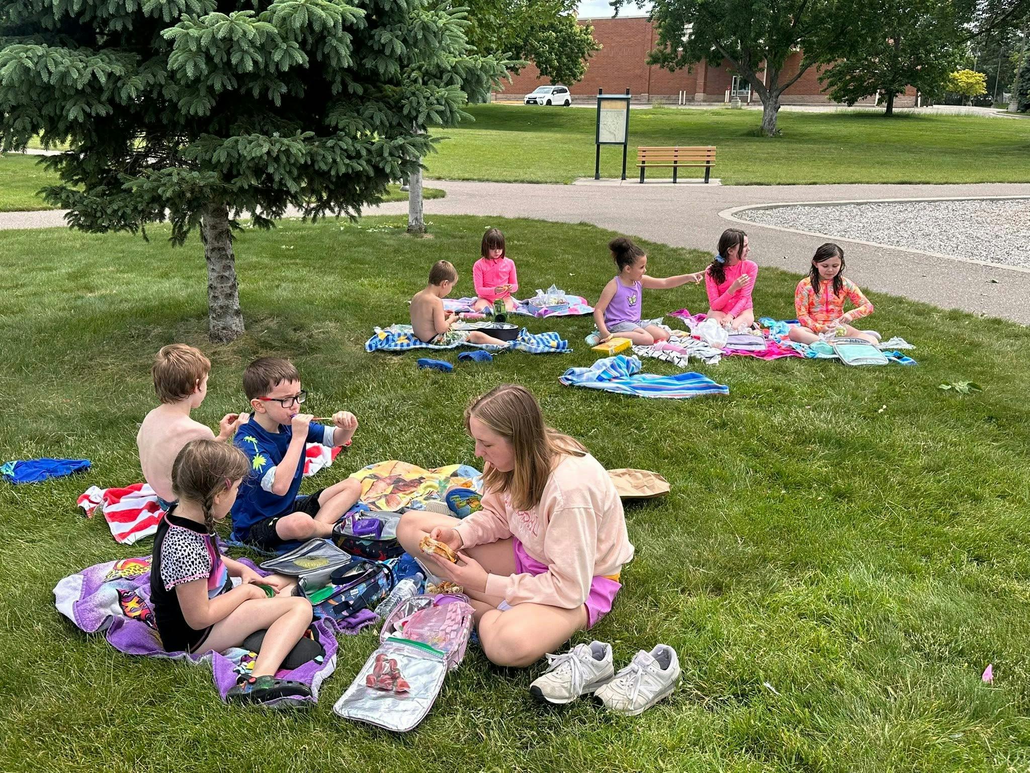 Group of children and an adult eating lunch on towels in a park