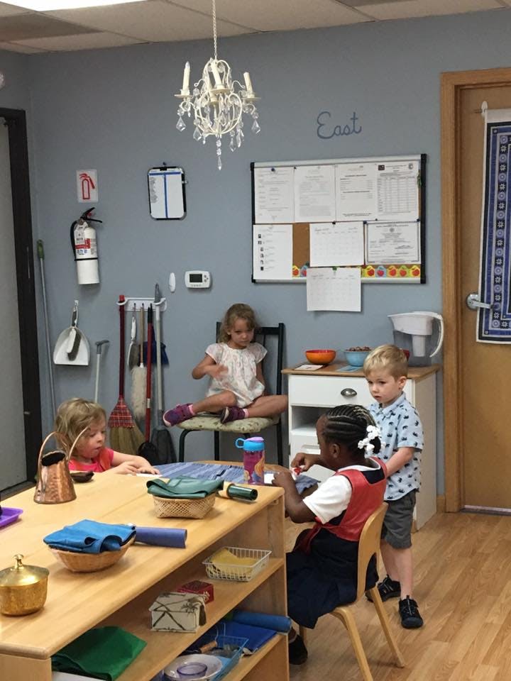 Children in a classroom, one sitting on a chair, one at a table, one standing