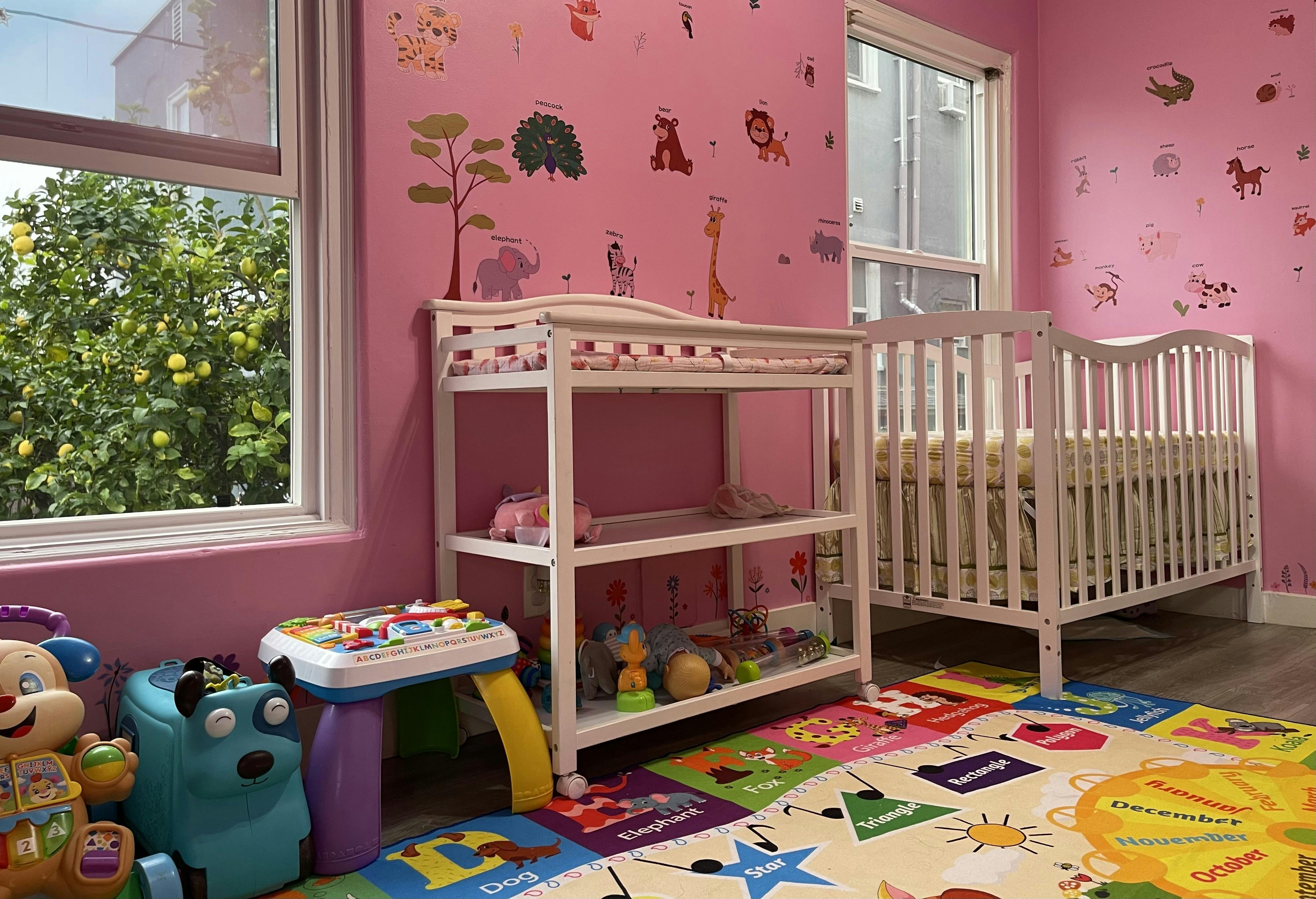 A pink nursery room with animal wall decals, a white crib, changing table, and play mat