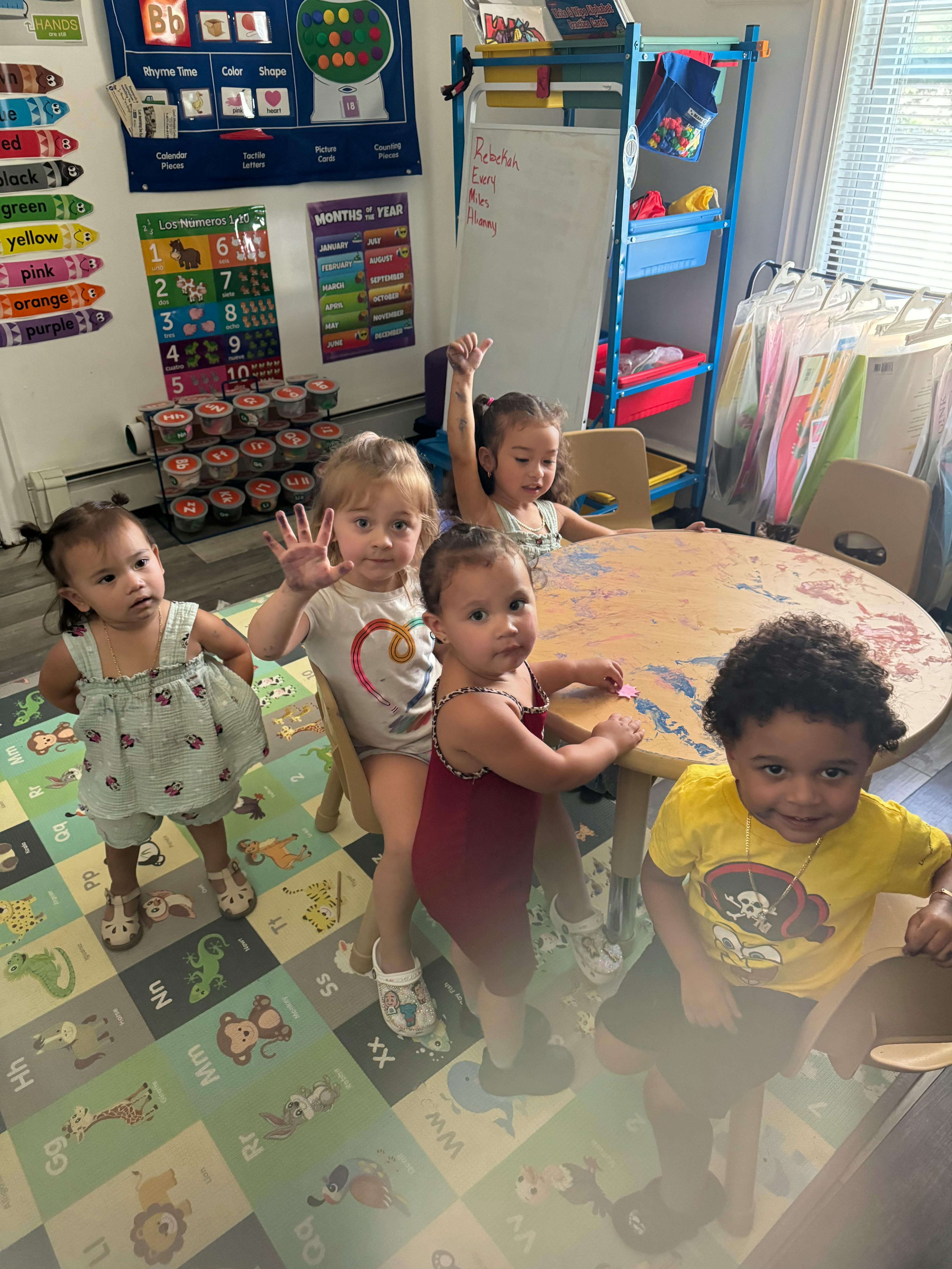 Five children in a classroom, some standing, some sitting at a table