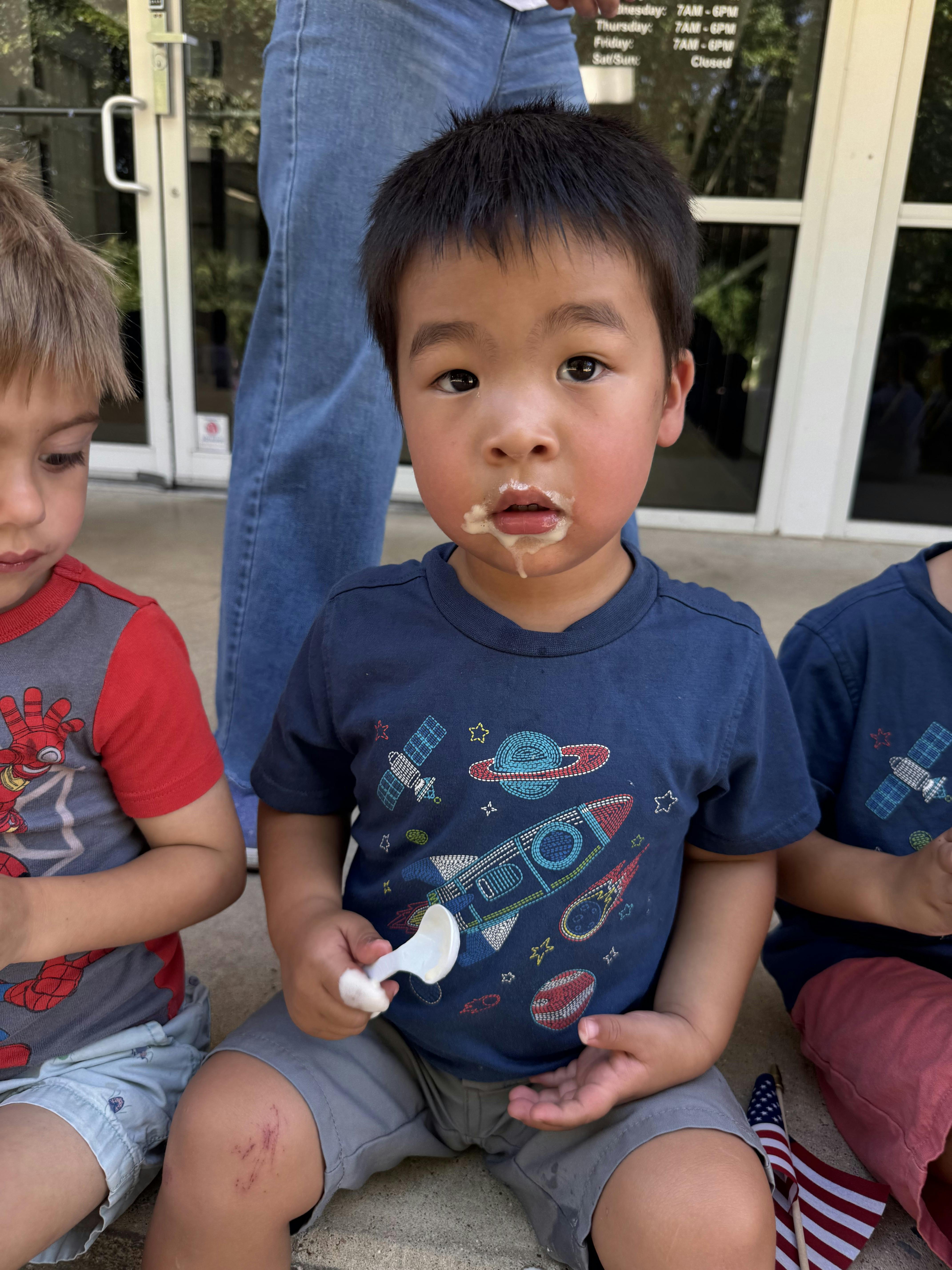Child with food on their mouth holding a spoon while sitting outdoors